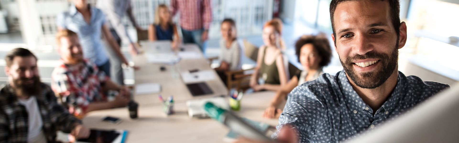 A man in the foreground smiling while writing something just below frame with a group of people watching in the background