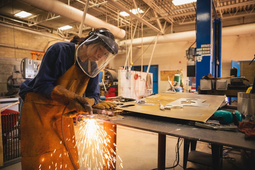 A person wearing a face shield and a leather apron while welding with sparks flying toward the ground